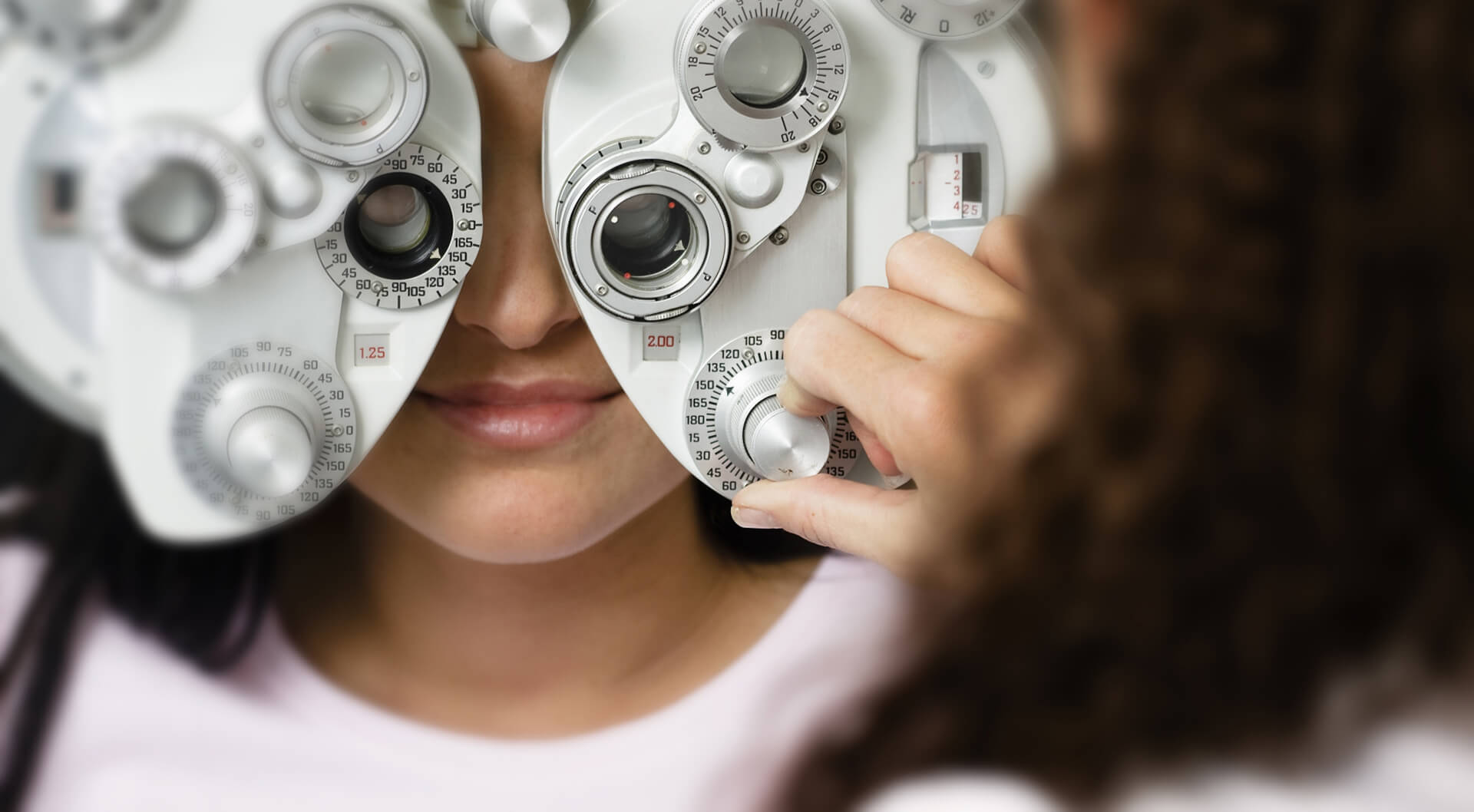 A close up shot of a person getting their eyes checked on a machine, with an optician adjusting knobs on the machine