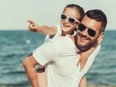 Happy little daughter in sunglasses sits on fathers back. Family on beach.