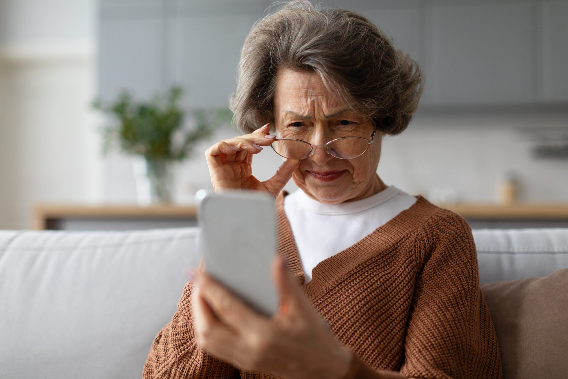 Older woman in her living room squinting at her phone screen and adjusting her glasses to see better.