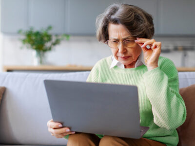 Older woman holding her glasses and squinting while using her laptop in her living room